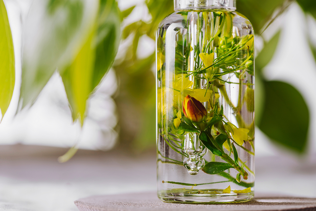 Glass bottle containing fresh botanical flowers and herbs infused in clear oil, representing ORY Skincare’s botanical infusion method designed to support every skin type through gentle, plant-based formulation.